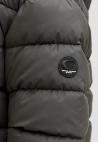 Czarna pikowana kurtka o błyszczącej fakturze, z okrągłą naszywką z logo na rękawie. Wzór zawiera poziome pikowane sekcje.