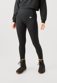 Svarte høye leggings med en glatt tekstur og en hvit logo foran, sammen med svarte og gull treningssko.