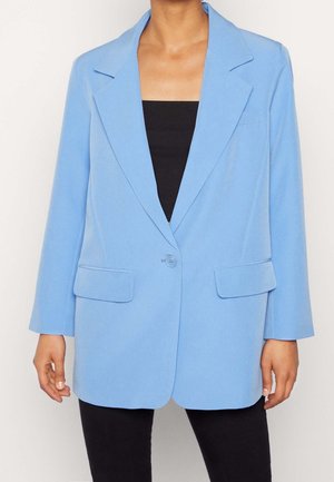 Femme portant un blazer bleu clair à bouton unique avec des revers crantés et des poches à rabat, par-dessus un haut noir et un pantalon noir.