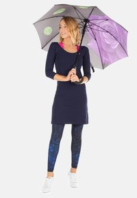 Abito blu navy con maniche lunghe, abbinato a leggings blu con un motivo astratto. Tieni un ombrello nero con disegni di foglie verdi e rose viola.