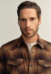 Man met kort bruin haar en baard, draagt een bruin geruit shirt over een wit T-shirt, kijkt recht vooruit met een neutrale uitdrukking.
