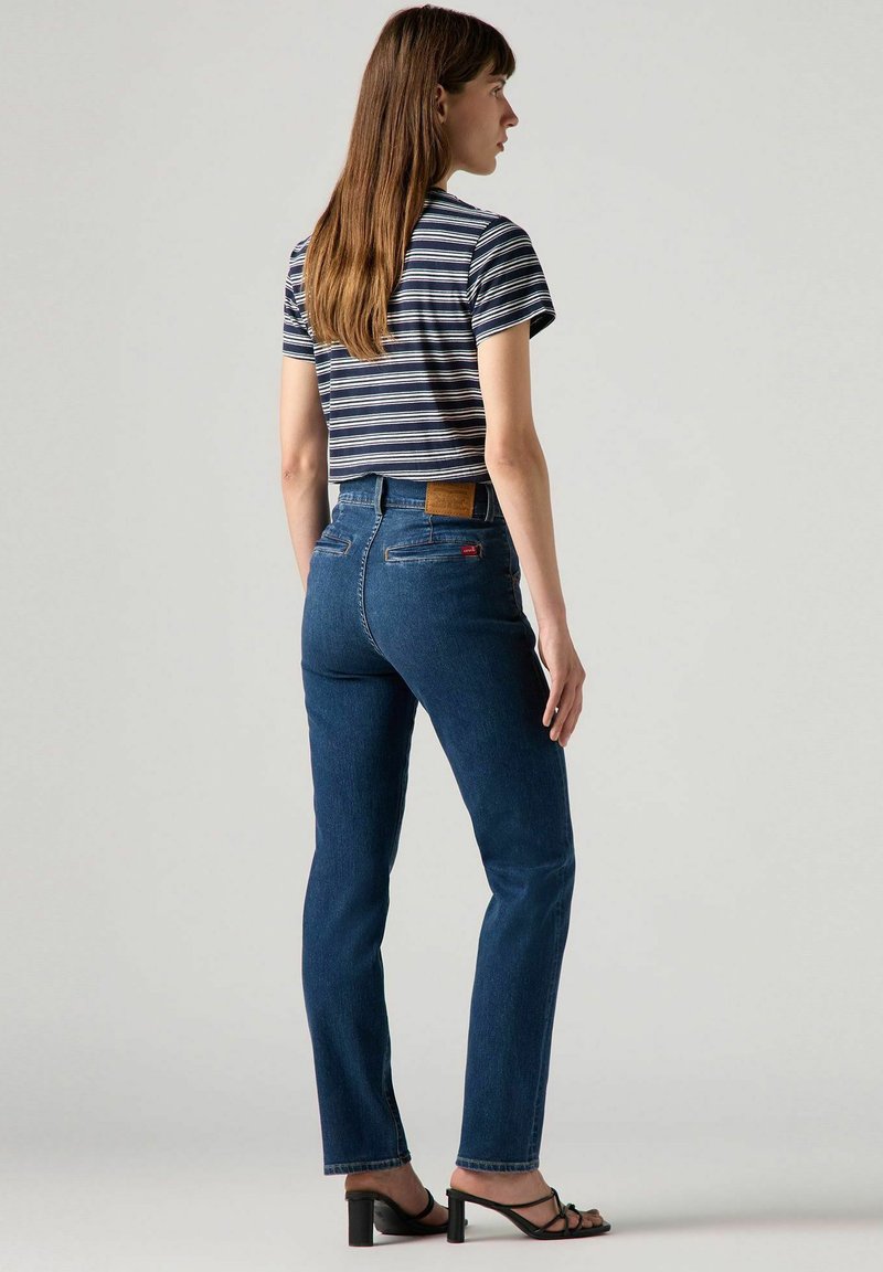 Jeans o wysokim stanie, proste nogawki w ciemnoniebieskim dżinsie z brązową skórzaną łatą z tyłu, w połączeniu z krótkim, paskowanym t-shirtem.