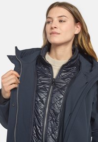 Marineblauer Blazer mit einem gesteppten Innenfutter. Verfügt über einen hohen Kragen, einen Frontreißverschluss und Druckknöpfe. Weiche Textur, geschichtetes Design.