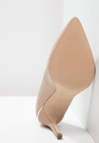 Chaussure à talon haut en cuir nude avec un bout pointu et un talon fin. La semelle présente des nervures horizontales pour une meilleure adhérence. Accent doré sur le talon.