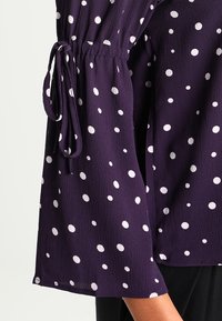 Blusa viola in tessuto leggero con pois bianchi, con maniche ampie strette ai polsi da nastri. Dettaglio della superficie strutturata.