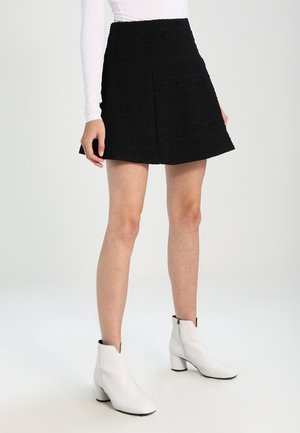 Femme portant une mini-jupe noire texturée en forme trapèze, un haut blanc à manches longues et des bottines blanches à talons blocs sur fond blanc.