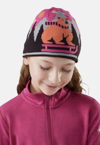 Ragazzina che indossa una giacca rosa con zip e un berretto lavorato a maglia colorato con un orso, su una slitta e un design di montagna, guarda in basso e sorride dolcemente.