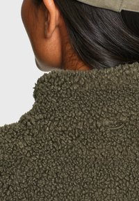 Gros plan sur le cou et l'oreille d'une personne portant une veste polaire vert olive avec une texture bouclée et une casquette beige.