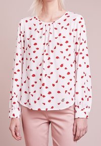 Blouse blanche à manches longues avec motif de feuilles rouges et grises, portée avec un pantalon rose pâle, montrée du cou aux hanches sur un fond rose doux.