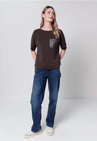 Frau steht in einem lässigen dunkelbraunen Oberteil mit Leopardentasche, blauen Jeans und weißen Sneakern vor schlichtem Hintergrund.