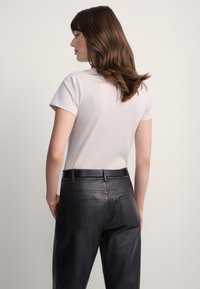 Kobieta o brązowych włosach do ramion, ubrana w jasną bluzkę z krótkim rękawem, wpuszczoną do wysokich czarnych skórzanych spodni z tylnymi kieszeniami.