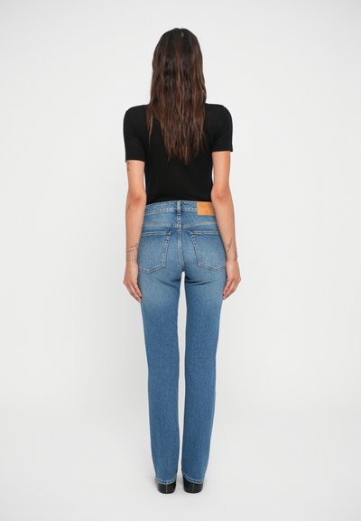 Jeans de denim azul con corte recto, cintura alta y una etiqueta de marca de cuero marrón en la parte trasera. Textura suave con dos bolsillos traseros.