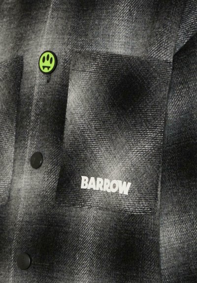 Zwart en grijs geruit flanellen shirt met een voorzak, zwarte knopen en een witte "BARROW" logo. Bevat een klein groen glimlach knoopdetail.