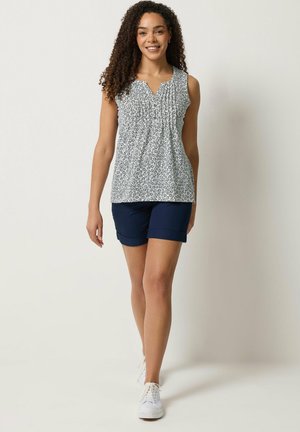 Mujer con cabello rizado, vestida con una blusa sin mangas estampada, pantalones cortos azul marino y zapatillas blancas, caminando hacia adelante sobre un fondo liso.