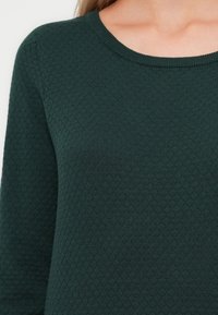 Mörkgrön stickad tröja med ett honungsfcomb-mönster, med rund nacke och långa ärmar, vilket ger ett mjukt och varmt intryck.