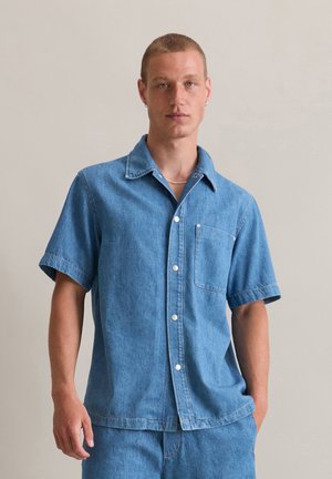 Lichtblauwe spijkerblouse met korte mouwen, knoopsluiting aan de voorkant, één borstzak, contrasterende stiksels, ontspannen pasvorm, textuurstof.