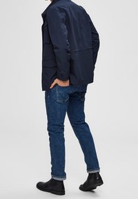 Marinblå jacka med framfickor, blå denimjeans och svarta ankelskor. Jeans är smala med uppvikt nederkant.