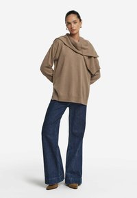 Beige trui met een gedrapeerde halslijn, gecombineerd met wijde, donkerblauwe jeans en bruine enkellaarzen. Glad textuur en losse pasvorm.