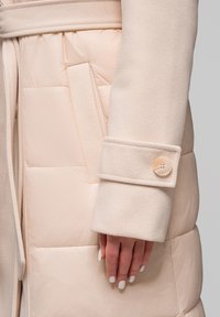 Detailní záběr na ruku s bílým lakem na nehty opřenou o béžový prošívaný kabát s vlněným manžetovým rukávem a detailem knoflíku.