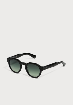 Lunettes de soleil noires à monture ronde avec des verres dégradés vert foncé et des branches épaisses sur un fond clair uni.