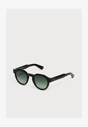 Lunettes de soleil noires à monture ronde avec des verres dégradés vert foncé et des branches épaisses sur un fond clair uni.