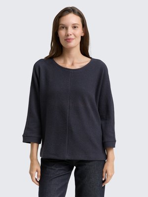Donker marineblauw langemouwen shirt met een gestructureerd patroon, voorzien van een brede ronde hals en opgerolde manchetten, gecombineerd met donkere jeans.