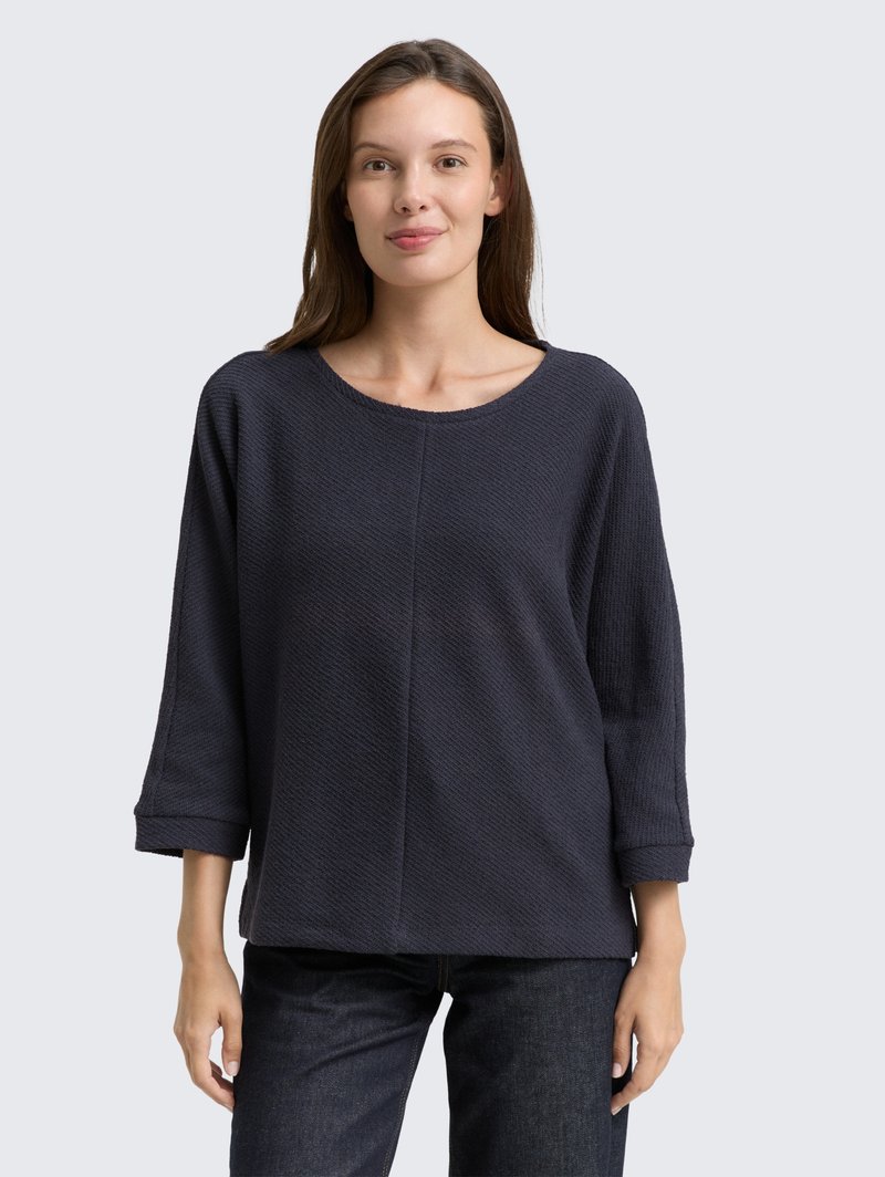 Dark navy long-sleeve top with a textured pattern, featuring a wide crew neckline and rolled cuffs, paired with dark jeans.