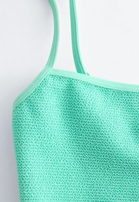 Gros plan d'un débardeur en tissu texturé vert menthe avec de fines bretelles, sur fond blanc.