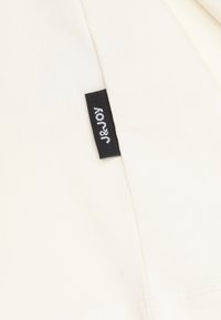 Crème-kleurige stof met een klein zwart label met de tekst "J&JOY". Gladde textuur met een subtiele glans, zonder zichtbare patronen of versieringen.