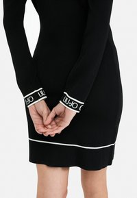 Černé pletené šaty s dlouhými rukávy, bílými akcentními lemy a logem na manžetách. Design má přiléhavý siluetu a rovný lem.