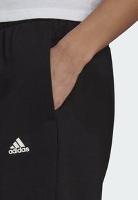 Svarta träningsbyxor med avslappnad passform, med en sidficka och en vit Adidas-logotyp. Tillverkade av mjukt, strukturerat material.