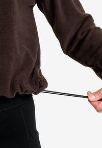Person, der den elastischen Kordelzug an der Taille der braunen Fleecejacke über schwarzen Hosen vor einem weißen Hintergrund anpasst.
