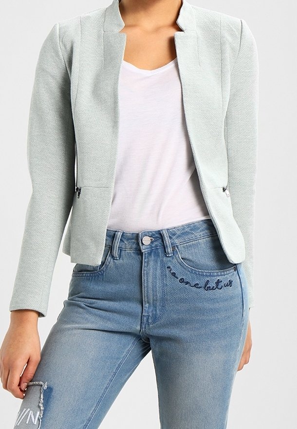 Blazer vert clair à finition texturée, à l'avant ouvert et sans col. Associé à un jean bleu clair avec un texte brodé sur la poche.