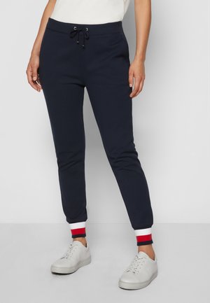 Personne portant un pantalon jogger bleu marine avec des poignets blancs, rouges et bleu marine, des baskets blanches et un haut blanc, debout devant un fond uni.