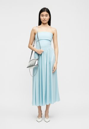 Mujer con un vestido midi azul claro sin tirantes, sosteniendo un bolso metálico plateado con cadena, y llevando zapatos de tacón blancos puntiagudos, de pie.