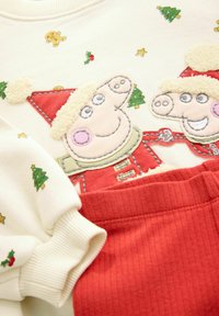 Hvid sweatshirt med et festligt broderet design af tegneseriegrise i nissehuer, med små juletræsmønstre. Røde ribbede bukser.