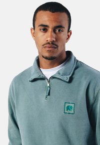 Sudadera de forro polar en verde azulado apagado, con cuello de media cremallera, logo de elefante bordado y tejido texturizado con un aspecto suave.