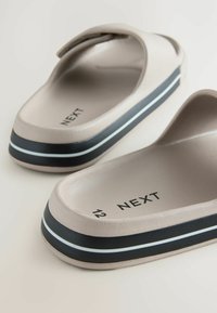 Nächste Marke Riemchensandalen aus beigem Gummi, mit einer strukturierten Oberfläche, schwarzer Sohle mit weißen Streifen und einem offenen Zehendesign.