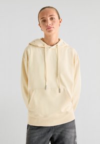 Beige hoodie met een trekkoordcapuchon, ribgebreide manchetten en zoom, een voorzak en een zachte textuur. Gecombineerd met donkergrijze jeans.