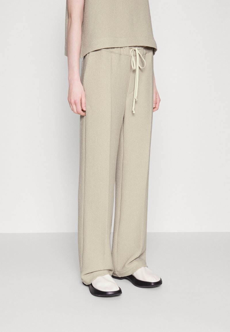 Personne portant un pantalon ample beige avec une taille à cordon, associé à des chaussures blanches avec des semelles noires, debout sur un sol clair.