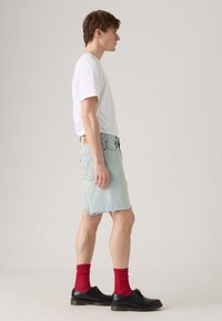 Shorts en denim bleu clair avec des bords effilochés, associés à un t-shirt blanc et des chaussettes rouges montantes. Chaussures en cuir noires à bouts ronds.