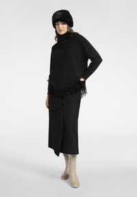 Schwarzer Pullover mit Fransen, Rollkragen, kombiniert mit einem schwarzen Rock und kniehohen beigen Stiefeln. Das Model trägt einen schwarzen Pelzhut.