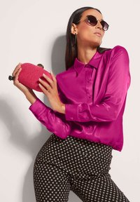 Fuchsia Satin-Bluse mit Kragen, kombiniert mit schwarzen Hosen mit weißen geometrischen Mustern. Hält eine strukturiert pinke Clutch-Tasche.