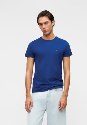 Young man with dark hair wearing a plain blue short-sleeve t-shirt and light blue jeans, standing against a plain background.