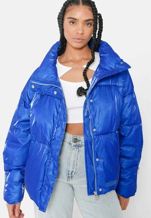 Frau mit geflochtenem Haar, die eine glänzende blaue Steppjacke über einem weißen, bauchfreien Top und hellblauen Jeans vor einem schlichten Hintergrund trägt.