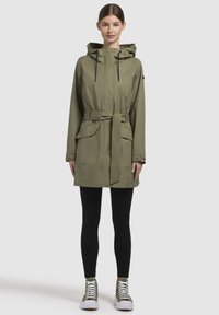 Olijfgroene waterdichte jas met capuchon, trekkoorden, ceintuur in de taille, twee voorzakken en een gladde textuur. Draagt over zwarte leggings en schoenen.