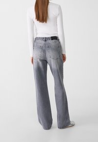 Grijze wijd gesneden jeans met een vervaagde textuur, vijf zakken, en een zwarte riemlus aan de achterkant, gecombineerd met een aansluitend wit shirt met lange mouwen.