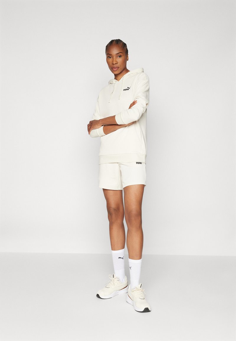 Hellbeige Kapuzenpullover mit Kordelzug, passende Shorts und weißen Socken. Mit kleinen schwarzen Logo-Akzenten; glattes Material, athletische Passform.