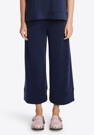 Femme portant un pantalon court large bleu marine et des chaussures à enfiler roses cloutées, debout devant un fond uni.