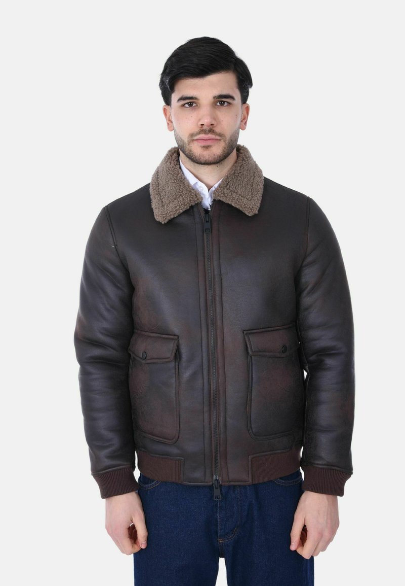 Giovane uomo che indossa una giacca di pelle marrone scuro con collo in shearling e tasche frontali, in piedi contro uno sfondo bianco semplice.